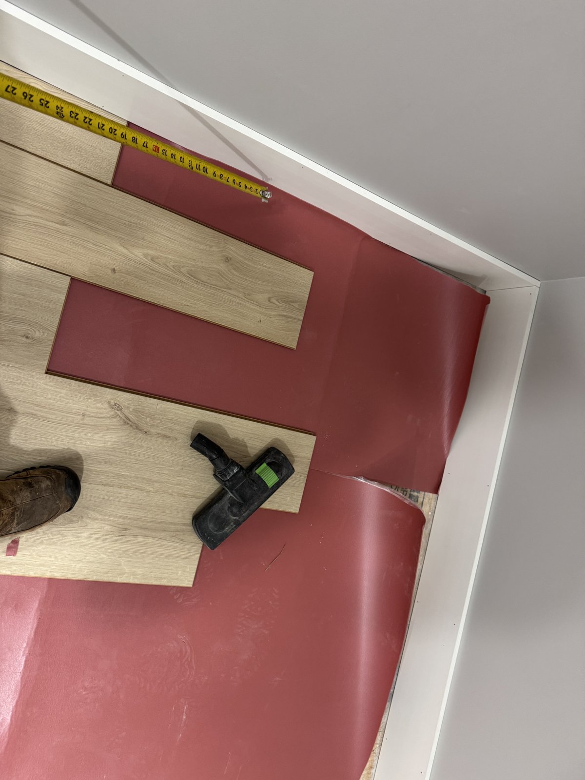 New plank flooring being laid over red underlayment in the finished attic, with a tape measure and vacuum on the floor